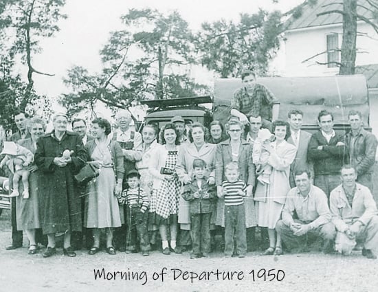 A group of men, women, and children standing in front of a loaded pickup truck with a farmhouse in the background