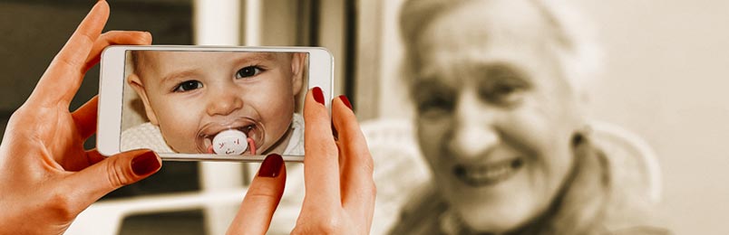 a photo of a smiling elderly woman facing a woman who is holding a phone with a photo of a baby on the screen.