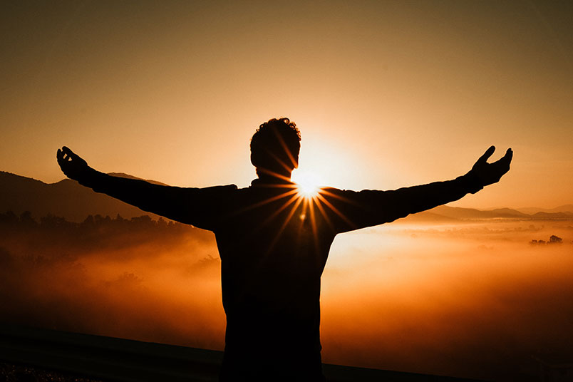 A photo of a man with arms outstretched facing a sunrise.