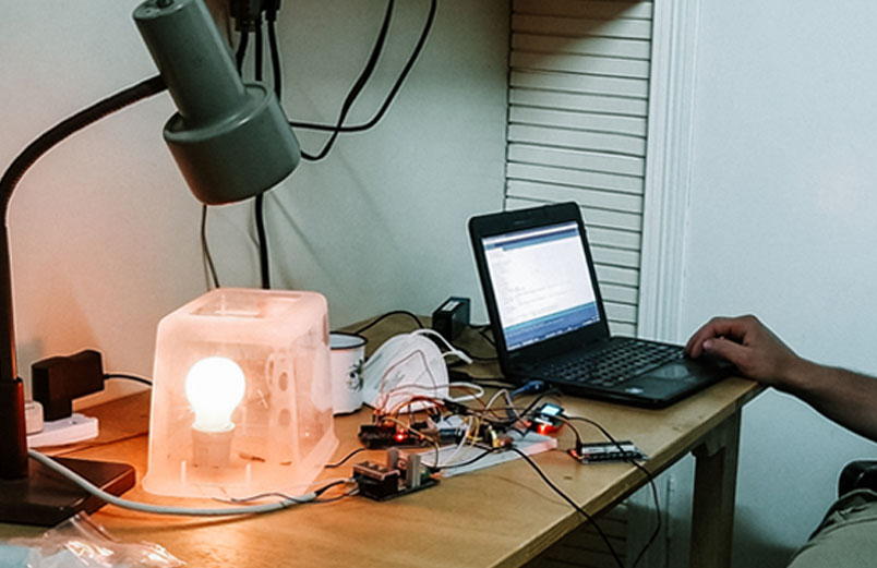 A man working at a computer beside circuit boards connected by wires and a light under a clear plastic box