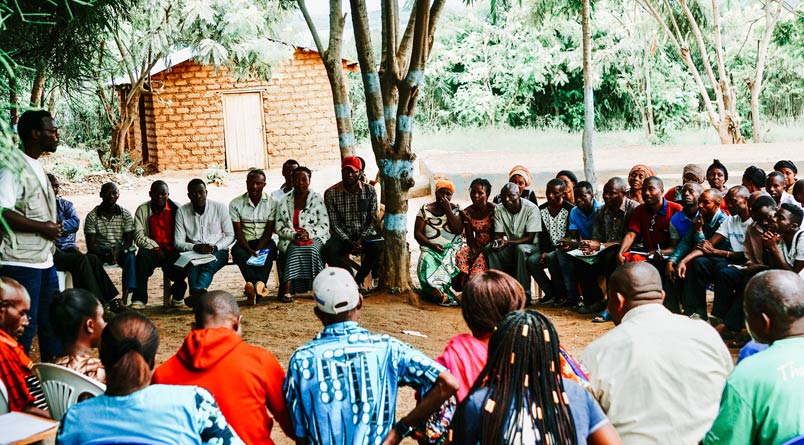 Men and women sitting outside in a circle on benches and in chairs listening to a standing man speaking to them.