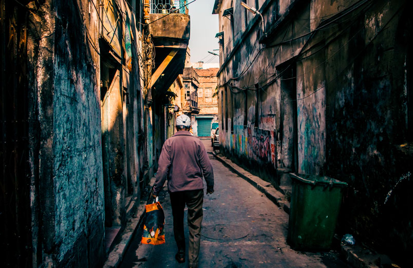 A man carrying a bag, walking down an alley in a city, away from the viewer. 