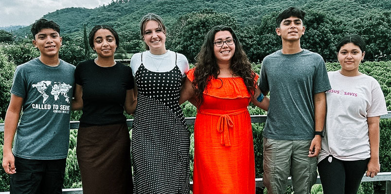 Six teens standing in a row on a blacony with mountains in the background