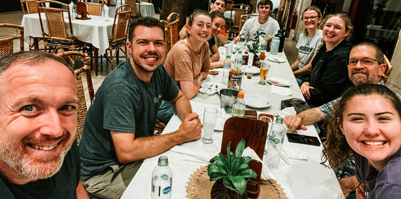 A group of people sitting around a table and smiling at the camrea