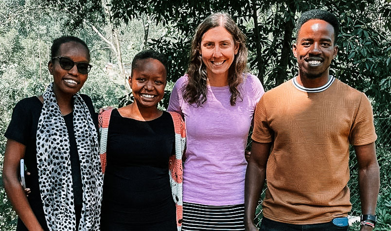 Three women and one man, standing side by side in front of some trees posing for a photo