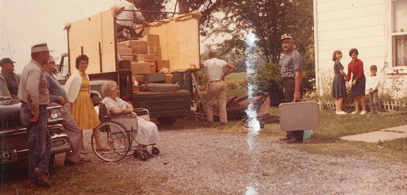 An old photo of a men packing the back of a pickup truck with a box built over the truck bed with family and friends looking on.