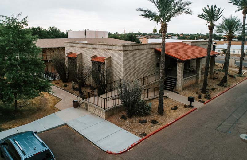 A one story building with a handicap accessible entrance and four palm trees in front on the corner of two streets.